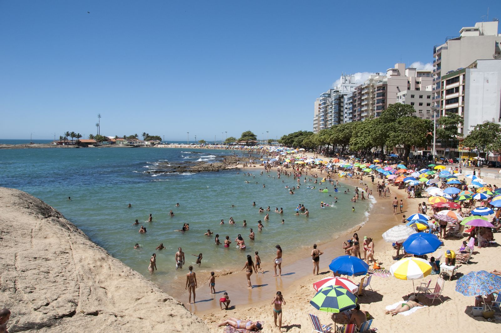 Praia das Castanheiras cheia de banhistas em um dia de sol, com guarda-sóis coloridos e prédios na orla