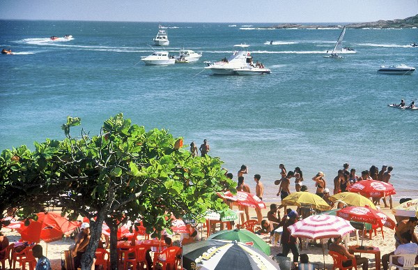 Praia da Bacutia movimentada, com banhistas, guarda-sóis e barcos ancorados no mar